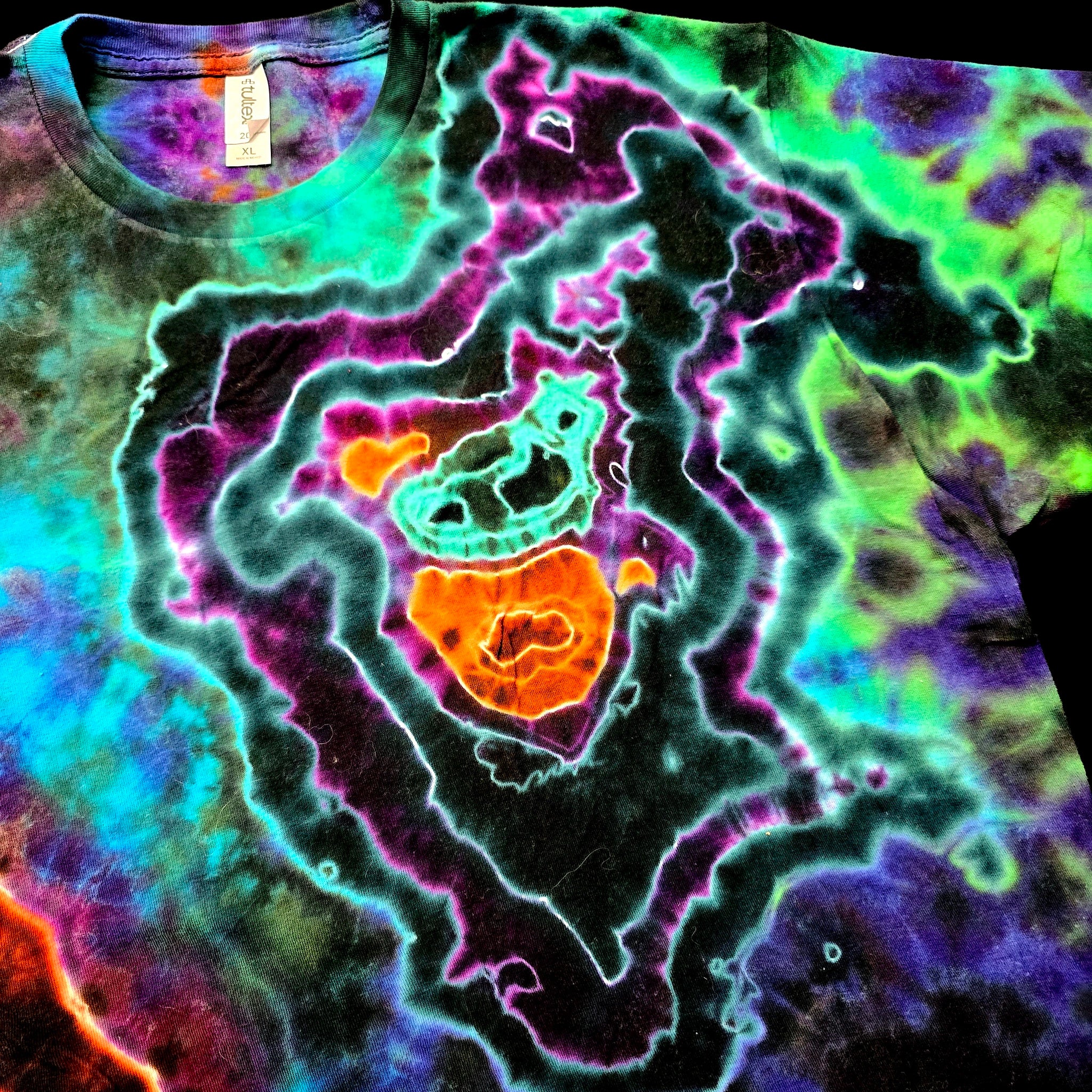 A close-up of a tie-dye t-shirt featuring a vibrant, multi-colored psychedelic pattern with concentric shapes in neon green, purple, orange, and blue over a black base.