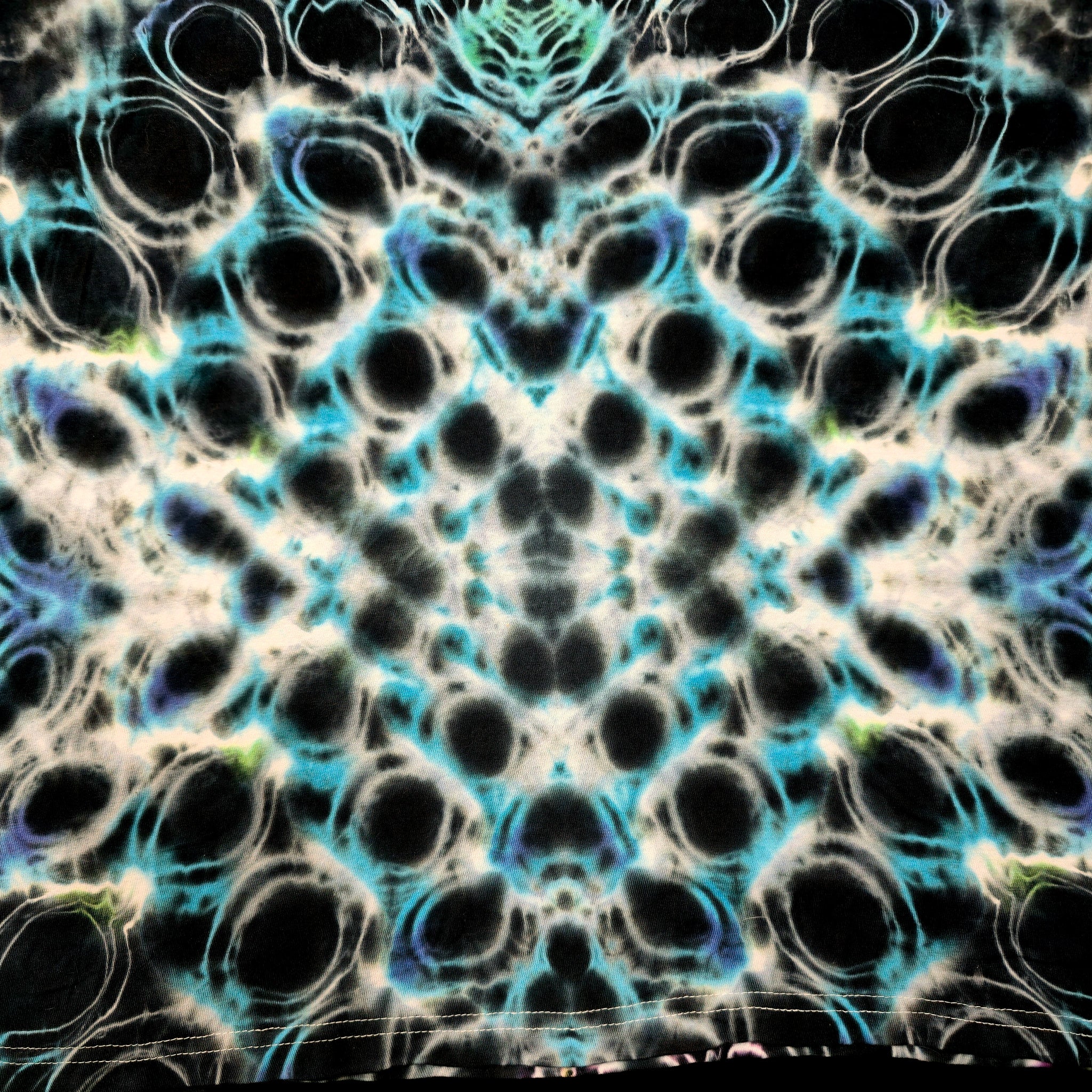 A close-up of a tie-dye fabric with a symmetrical, kaleidoscopic pattern featuring black, white, bright cyan blue, and subtle purple accents.