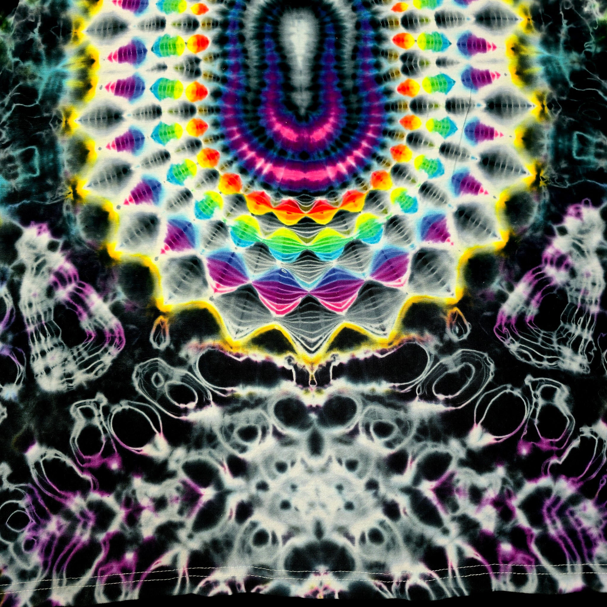 A highly detailed, symmetrical tie-dye pattern featuring a central multicolor mandala with concentric rings of pink, purple, blue, green, and yellow, surrounded by white and black wavy patterns.