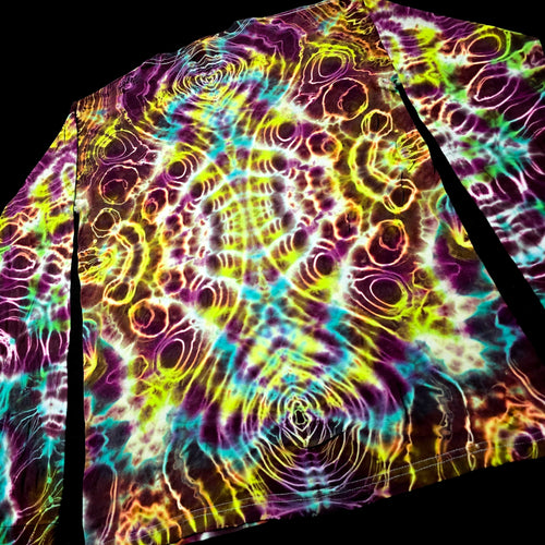 A close-up of a tie-dye patterned fabric, likely a shirt or towel, featuring a vibrant psychedelic design with concentric circles and wavy lines in purple, yellow, green, blue, and orange on a black background.