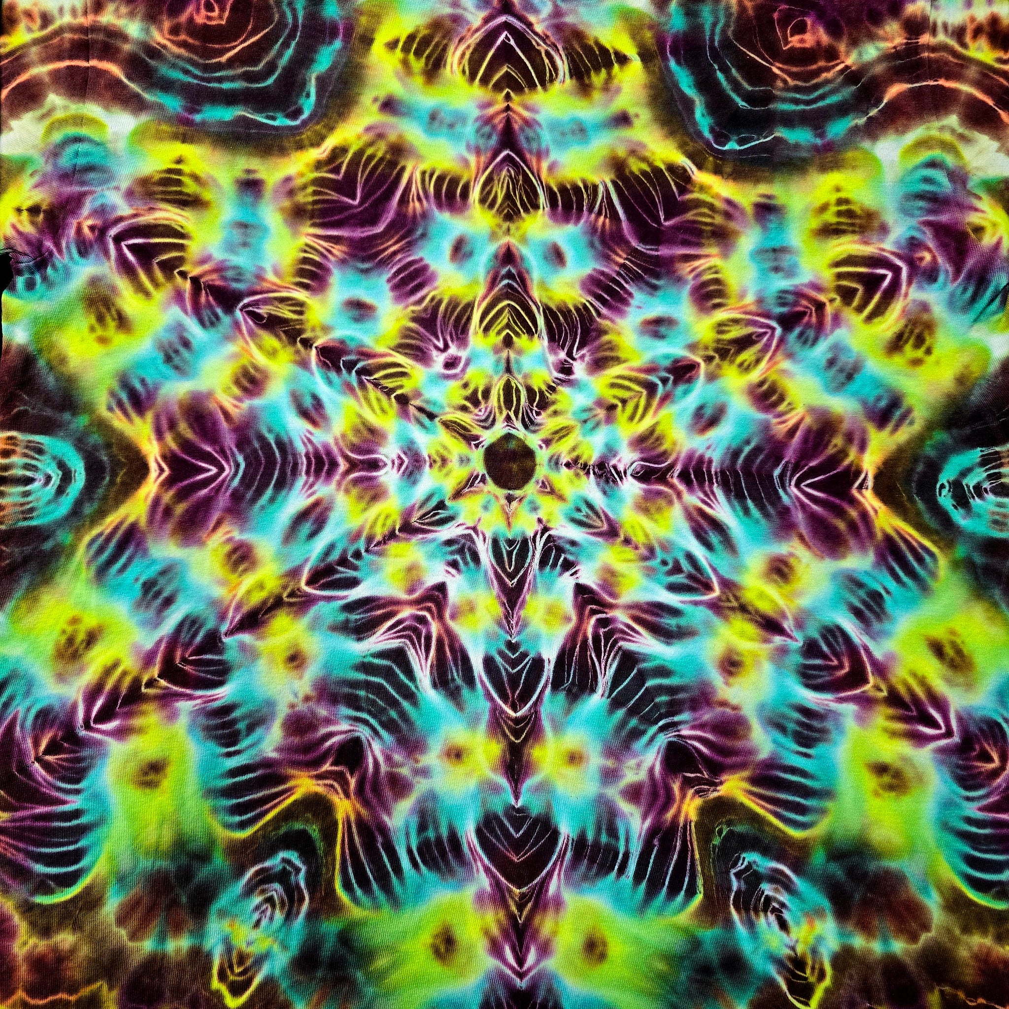 A square fabric or material swatch featuring a vibrant, symmetrical tie-dye pattern with neon yellow, cyan, purple, and dark brown/black tones arranged in a kaleidoscopic radial design.