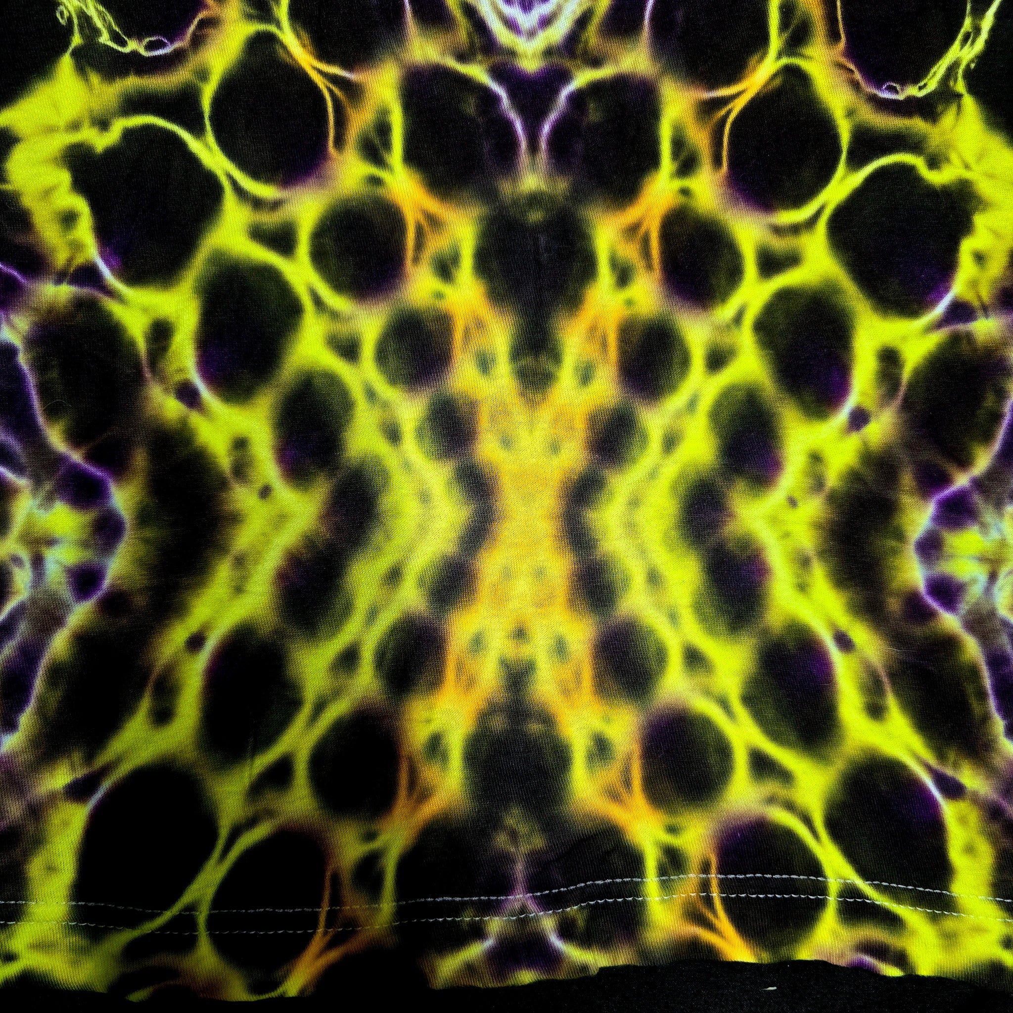 A close-up of a tie-dye fabric with a symmetrical, kaleidoscopic pattern in neon yellow, black, and purple, showing a stitched hem at the bottom.