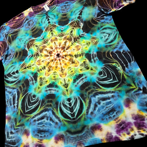 A short-sleeve tie-dye T-shirt laid flat on a black background, featuring a large symmetrical mandala-style starburst pattern in yellow, green, blue, purple, and black.