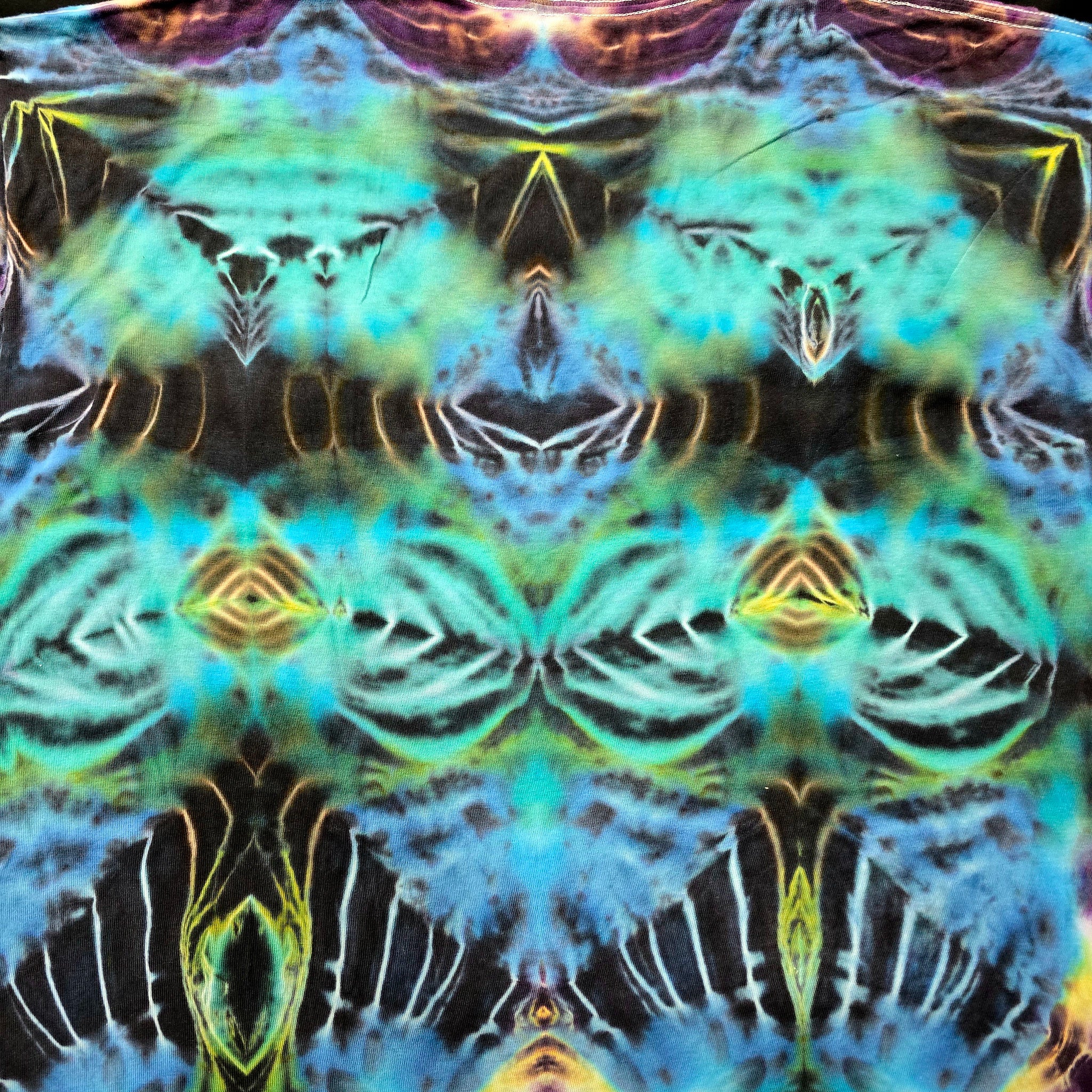 A close-up of a psychedelic tie-dye fabric with a symmetrical, kaleidoscopic pattern in vibrant shades of blue, green, yellow, and black.