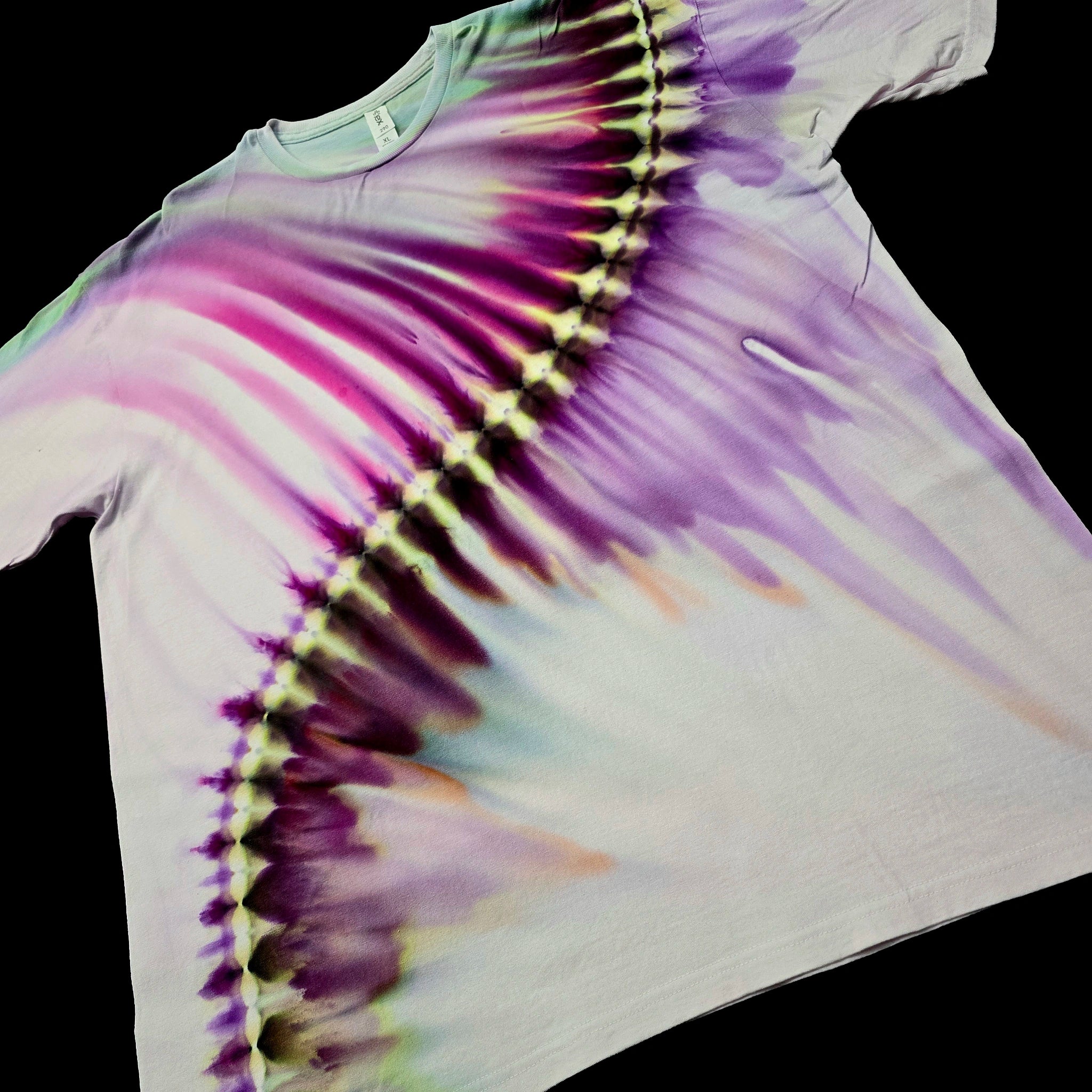 A white short-sleeve tie-dye t-shirt laid flat against a solid black background, featuring a diagonal rainbow tie-dye pattern with purple, pink, yellow, and green hues.
