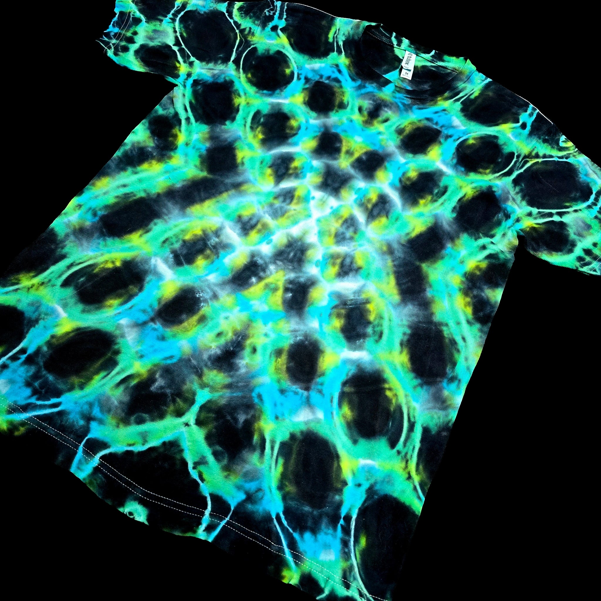 A short-sleeve crewneck t-shirt featuring a vibrant neon tie-dye pattern with glowing cyan, lime green, and electric blue cellular-like shapes over a black base, shown against a solid black background.