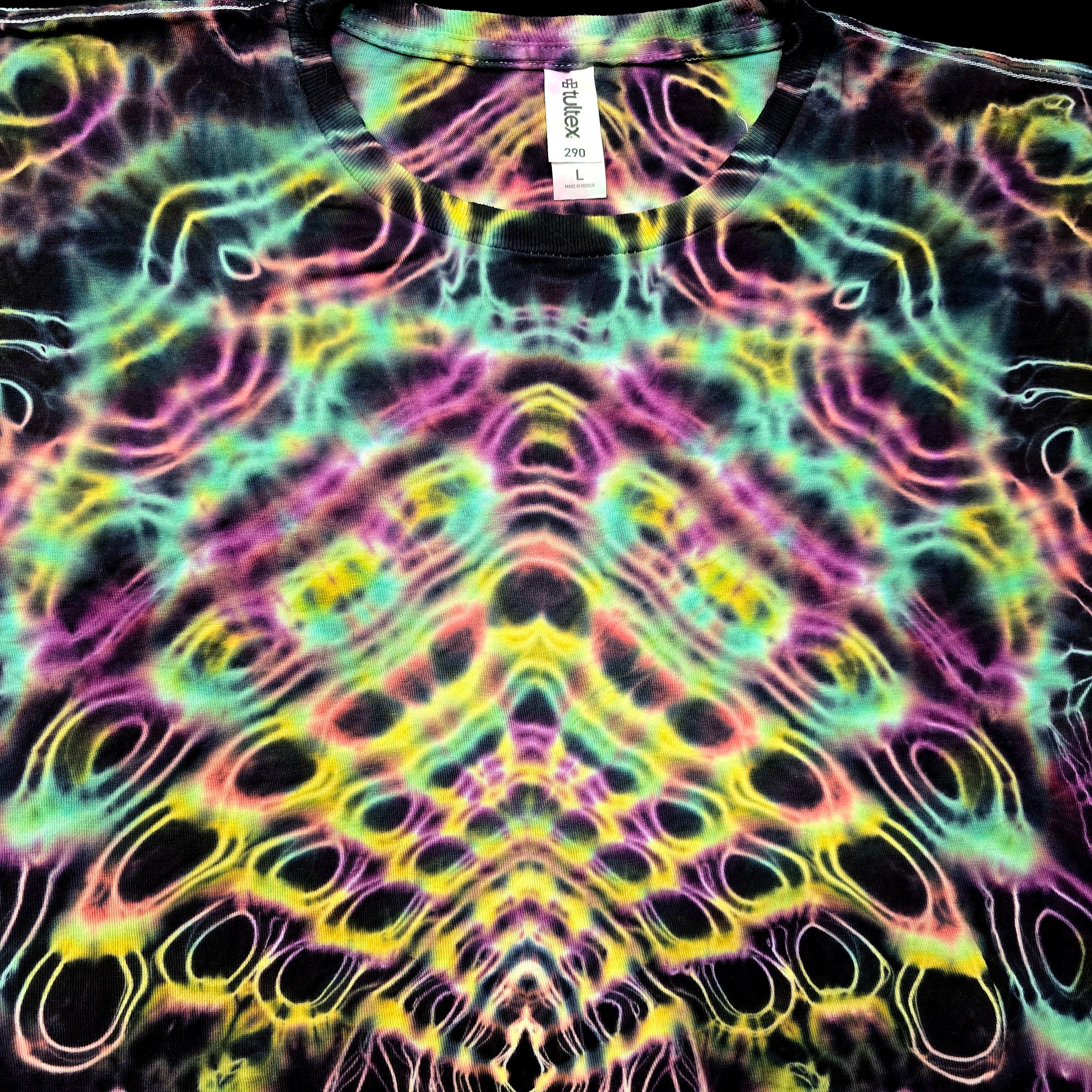 A close-up of a black t-shirt featuring a vibrant, symmetrical psychedelic tie-dye pattern with neon pink, green, yellow, and purple swirls. A white neck tag is visible at the collar.
