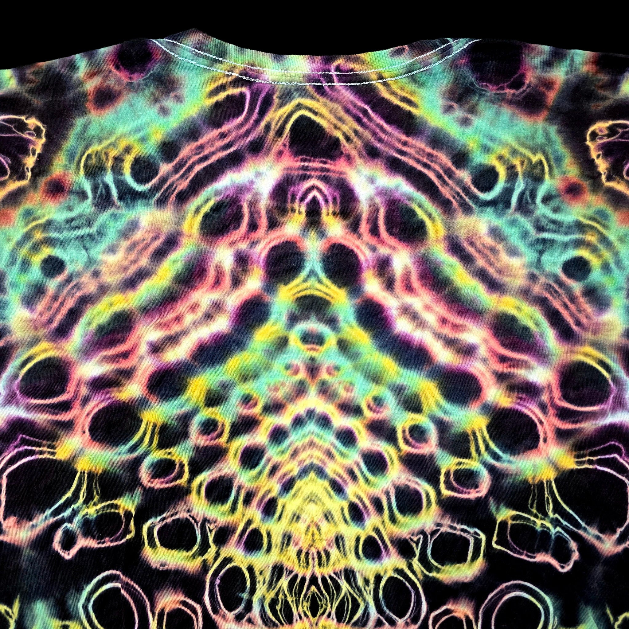 A close-up of a tie-dye t-shirt featuring a highly symmetrical, psychedelic pattern with neon pink, yellow, green, and blue outlines on a black base.