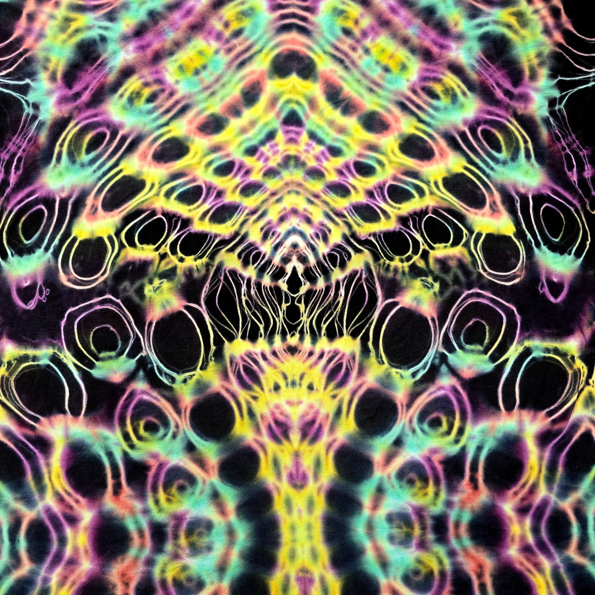 A close-up of a tie-dye fabric with a highly symmetrical, psychedelic pattern featuring neon yellow, pink, green, and purple concentric ring motifs on a black background.