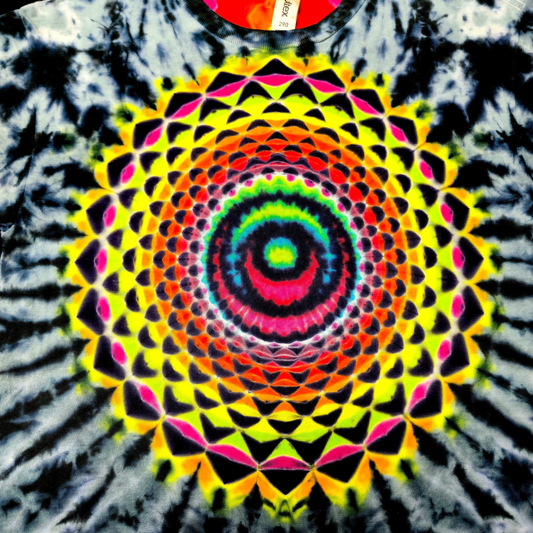 A close-up of a tie-dye t-shirt featuring a large, vibrant mandala pattern in the center with concentric circles and geometric shapes in neon yellow, green, blue, pink, red, and orange, surrounded by a black and white tie-dye burst.