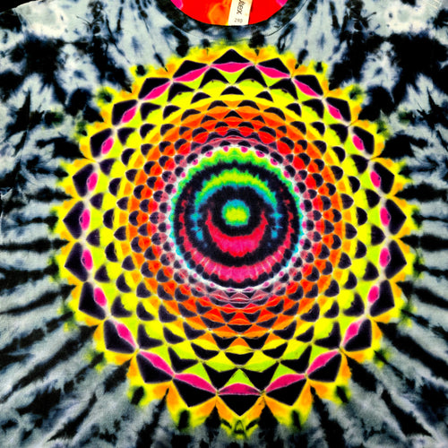 A close-up of a tie-dye t-shirt featuring a large, vibrant mandala pattern in the center with concentric circles and geometric shapes in neon yellow, green, blue, pink, red, and orange, surrounded by a black and white tie-dye burst.
