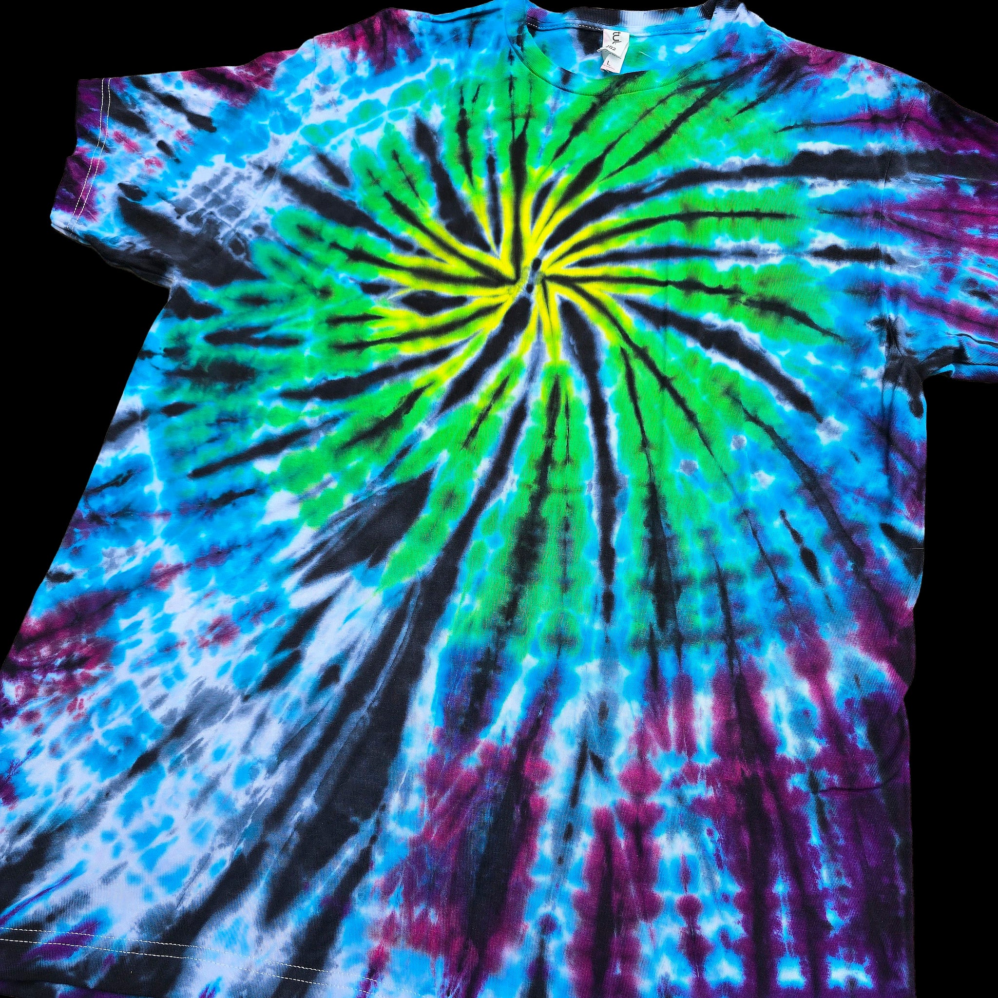 A short-sleeve tie-dye t-shirt featuring a vibrant spiral pattern with bright green, blue, purple, and black dye, laid flat against a black background.