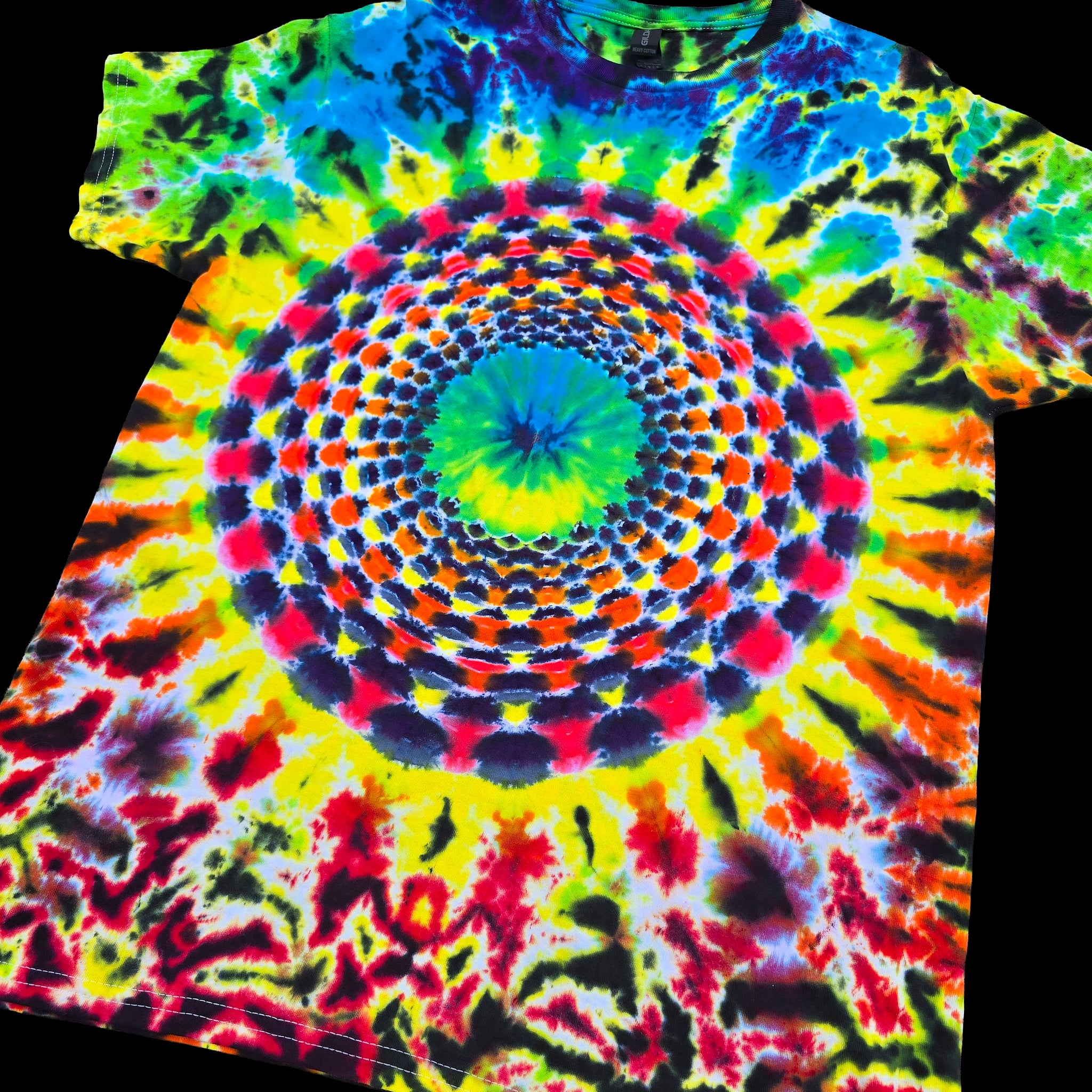 A short-sleeve tie-dye T-shirt laid flat on a black background, featuring a large concentric mandala-style spiral pattern in bright neon colors including yellow, green, blue, red, orange, purple, and black.