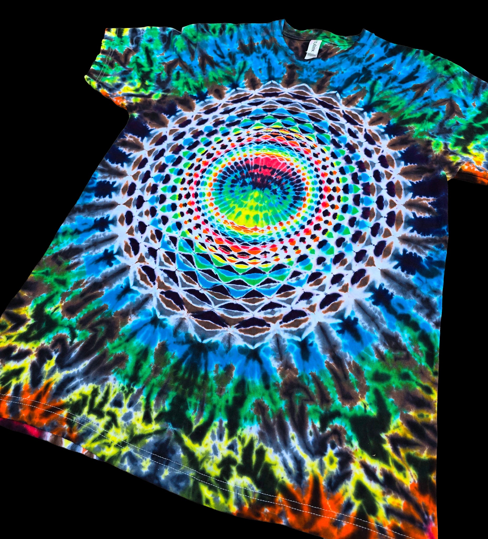 A short-sleeve tie-dye T-shirt laid flat on a black background, featuring a large circular mandala-style rainbow print with blue, green, yellow, red, orange, and white patterns.