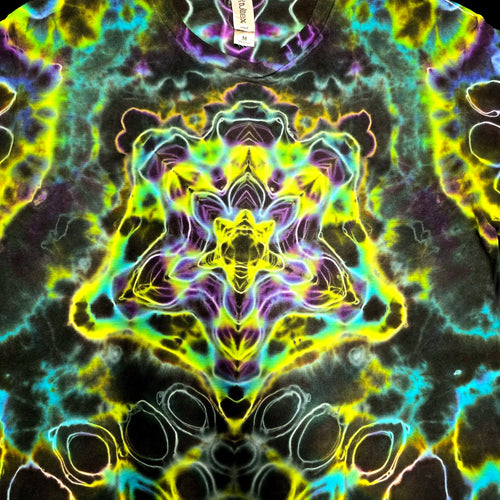 A close-up of a tie-dye garment featuring a vibrant, symmetrical psychedelic pattern with neon yellow, green, purple, and blue on a dark base. A small white paper tag is visible at the top center.