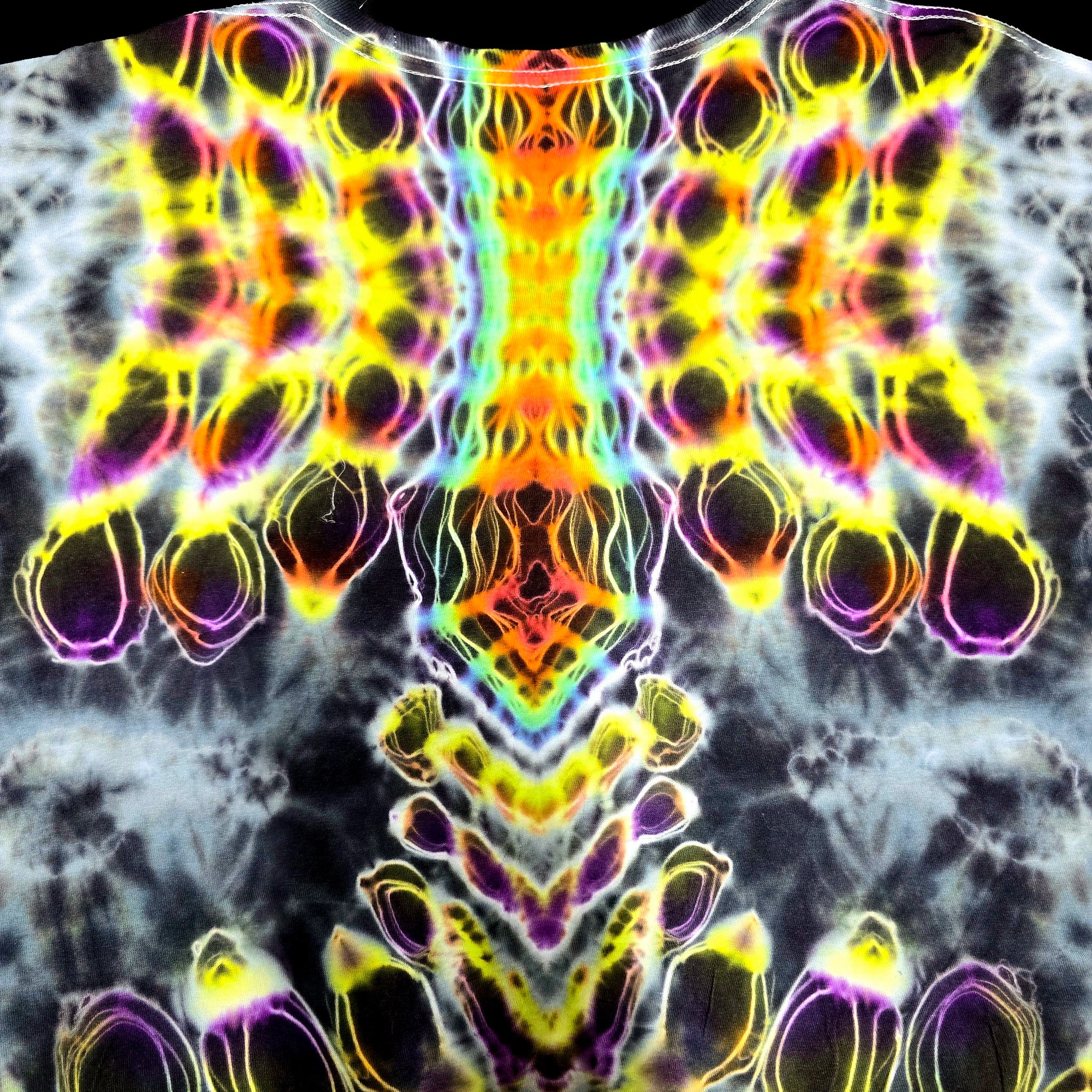 A close-up of a tie-dye garment featuring a highly symmetrical, psychedelic neon pattern with bright yellow, orange, green, blue, and purple swirls over a dark gray and white base.