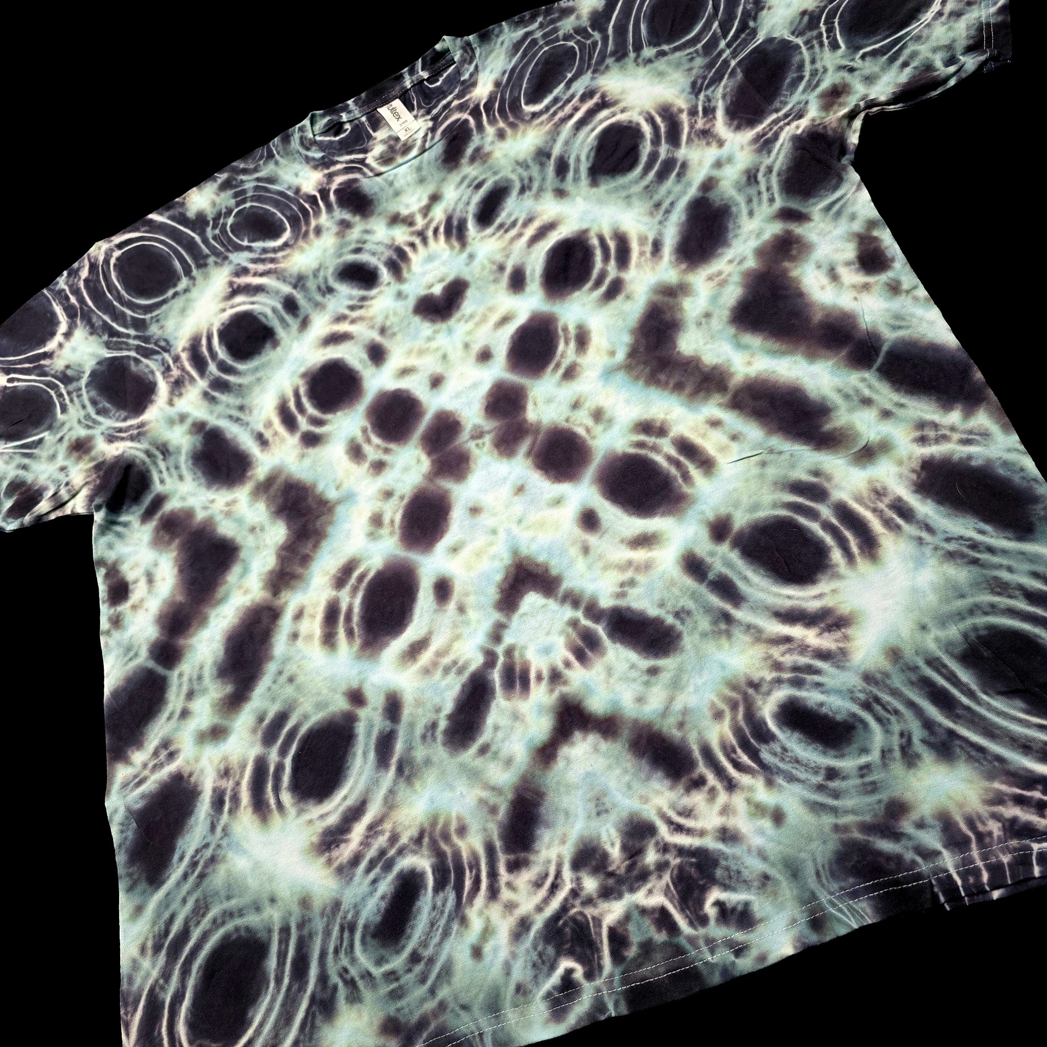 A short-sleeve t-shirt with an all-over psychedelic tie-dye pattern of concentric dark circles and wavy lines in black, dark brown, and off-white, shown against a solid black background.