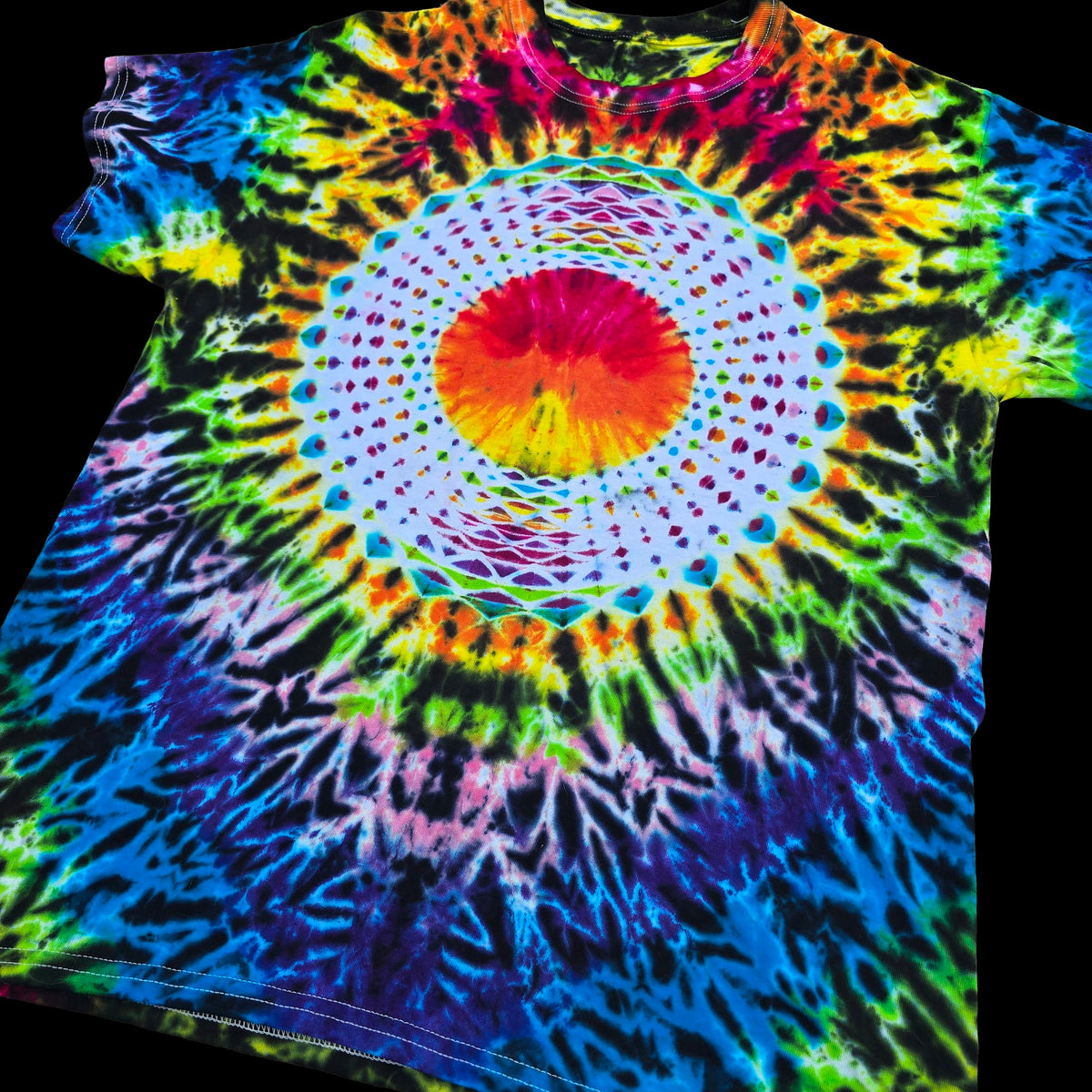 A short-sleeve tie-dye T-shirt laid flat against a black background, featuring a large circular mandala-style graphic in the center with concentric rings of red, orange, yellow, green, blue, purple, and pink fading into a rainbow tie-dye burst across the shirt.
