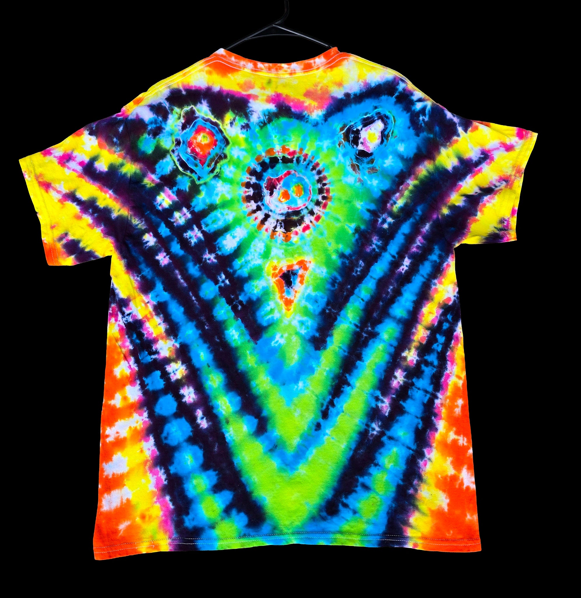 A short-sleeve tie-dye T-shirt hanging on a black hanger against a solid black background. The shirt features a highly symmetrical, psychedelic pattern with concentric circles and V-shaped bands of neon yellow, green, blue, purple, and orange.