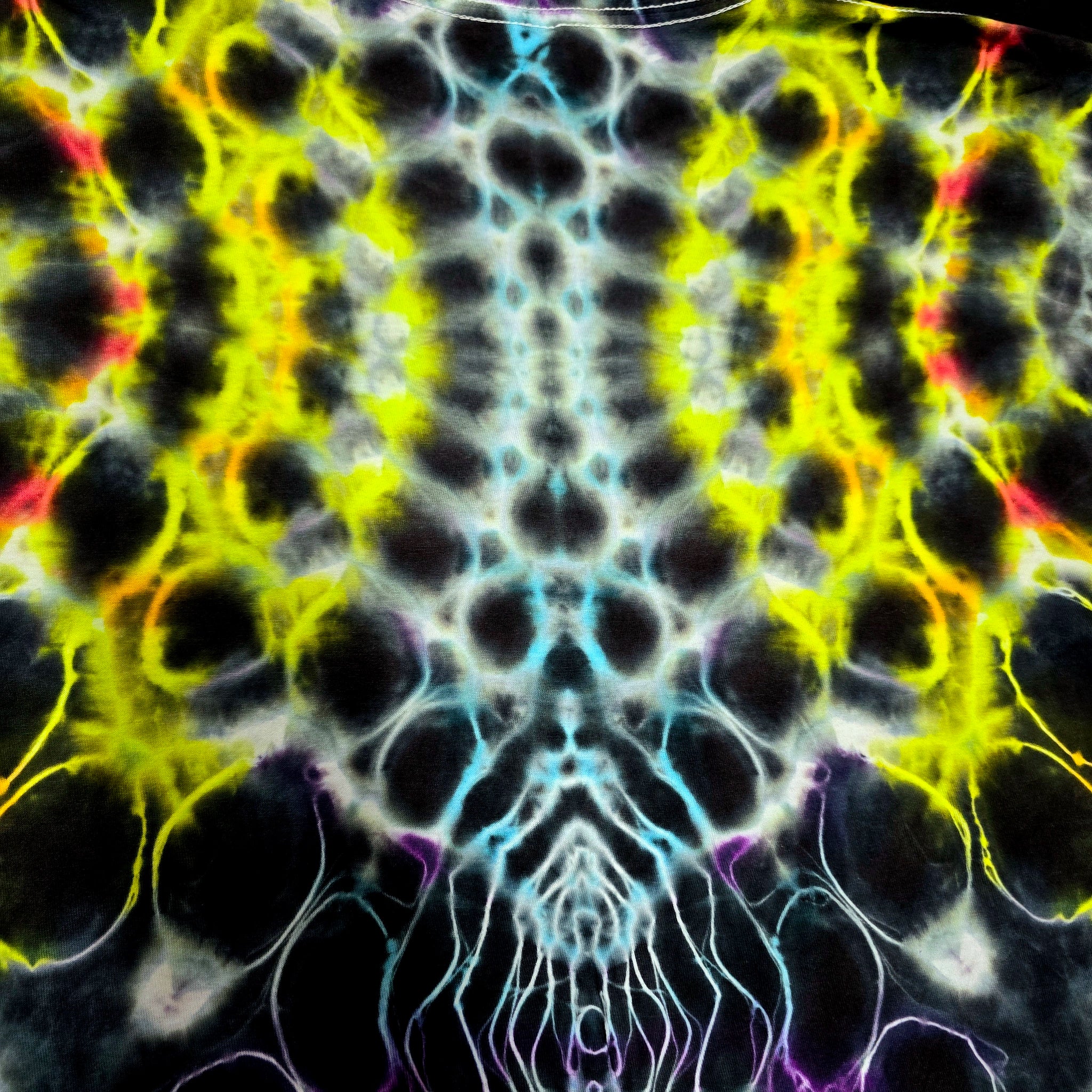 A close-up of a tie-dye fabric pattern with a symmetrical, kaleidoscopic design featuring black, neon yellow, light blue, red, and purple colors.