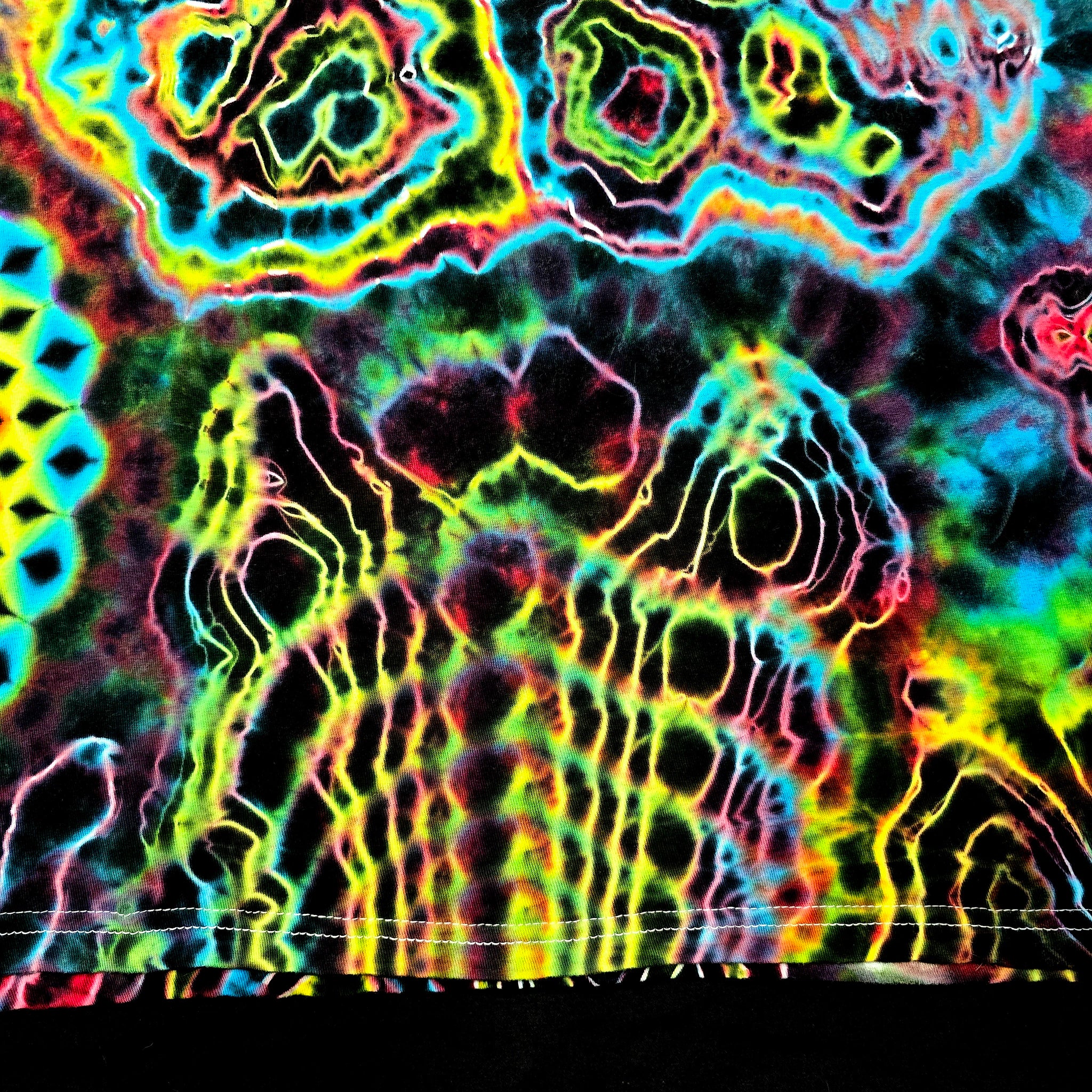 A close-up of a highly saturated tie-dye or psychedelic pattern with neon yellow, green, blue, pink, and purple outlines over a dark base. The fabric or material fills almost the entire frame, with a small black band visible along the bottom edge.