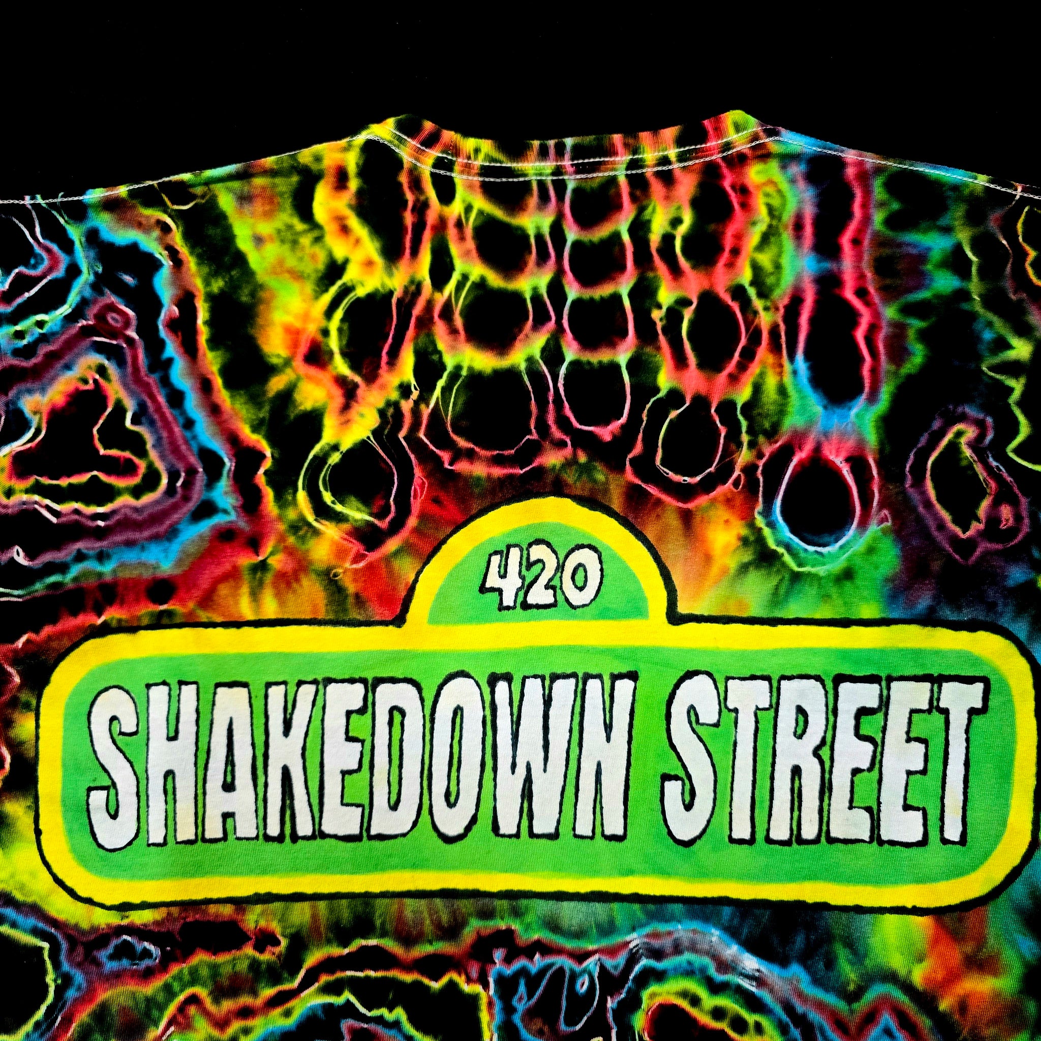 A close-up of a neon tie-dye t-shirt featuring a green street-sign style graphic that reads '420 SHAKEDOWN STREET' in white letters, set against a black background.