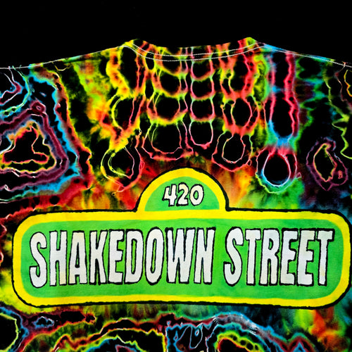 A close-up of a neon tie-dye t-shirt featuring a green street-sign style graphic that reads '420 SHAKEDOWN STREET' in white letters, set against a black background.