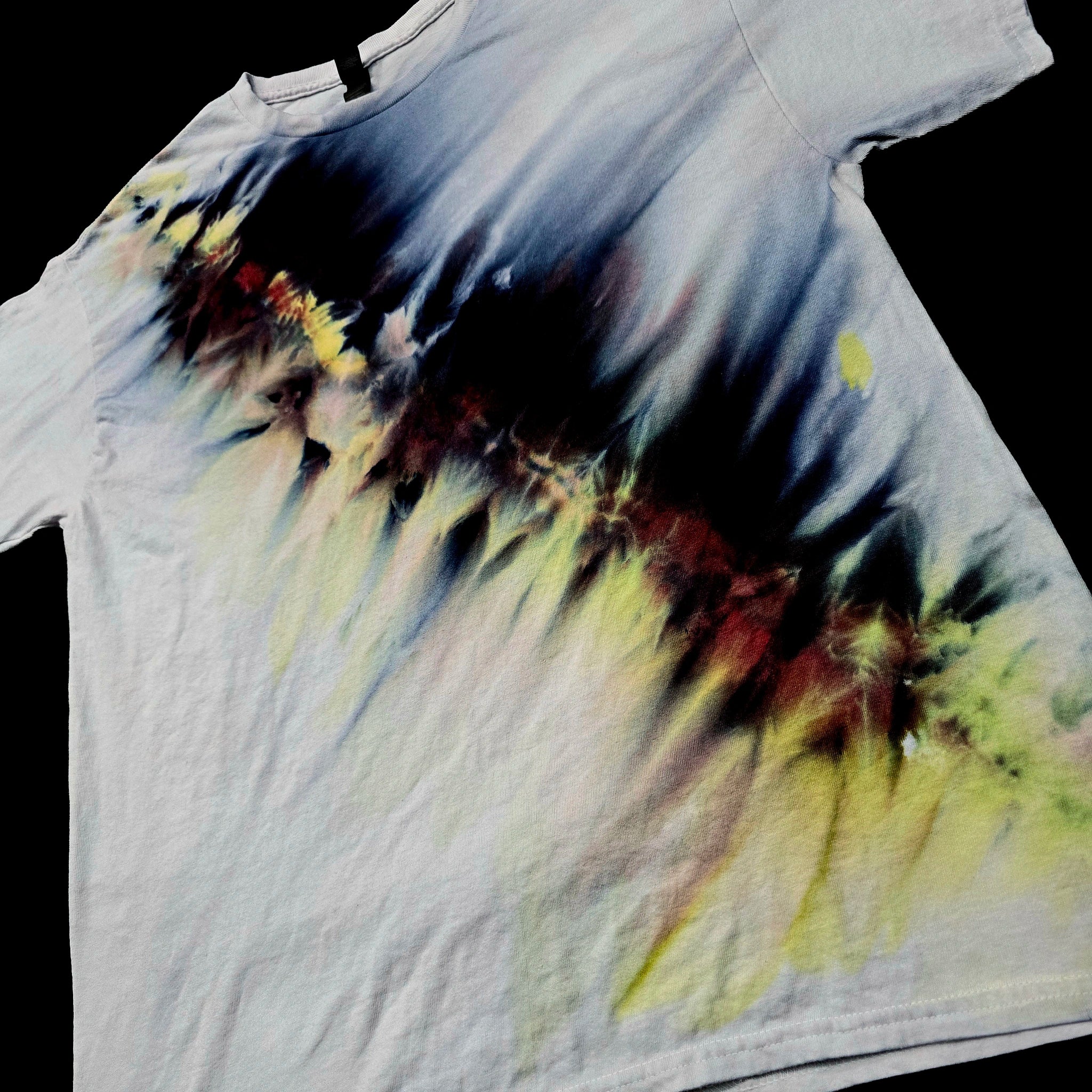 A white short-sleeve t-shirt with a dramatic tie-dye splash pattern in black, yellow, and red, laid flat against a solid black background.