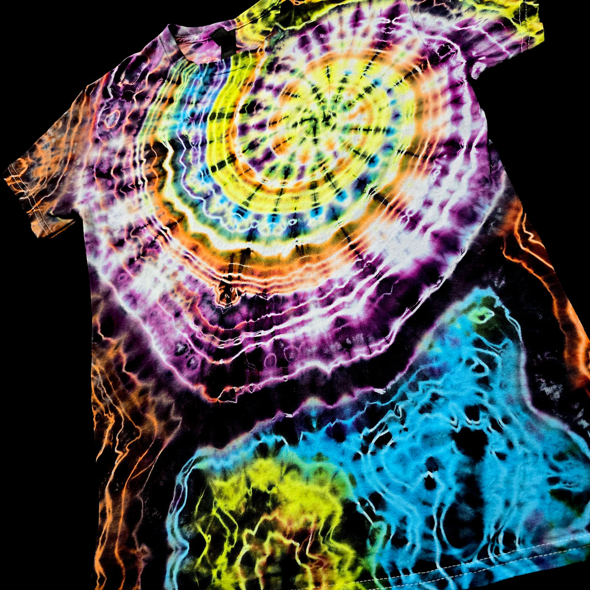 A short-sleeve tie-dye T-shirt with a large spiral mandala pattern in yellow, green, purple, pink, orange, and white, with a blue and yellow abstract section at the lower right, shown against a black background.