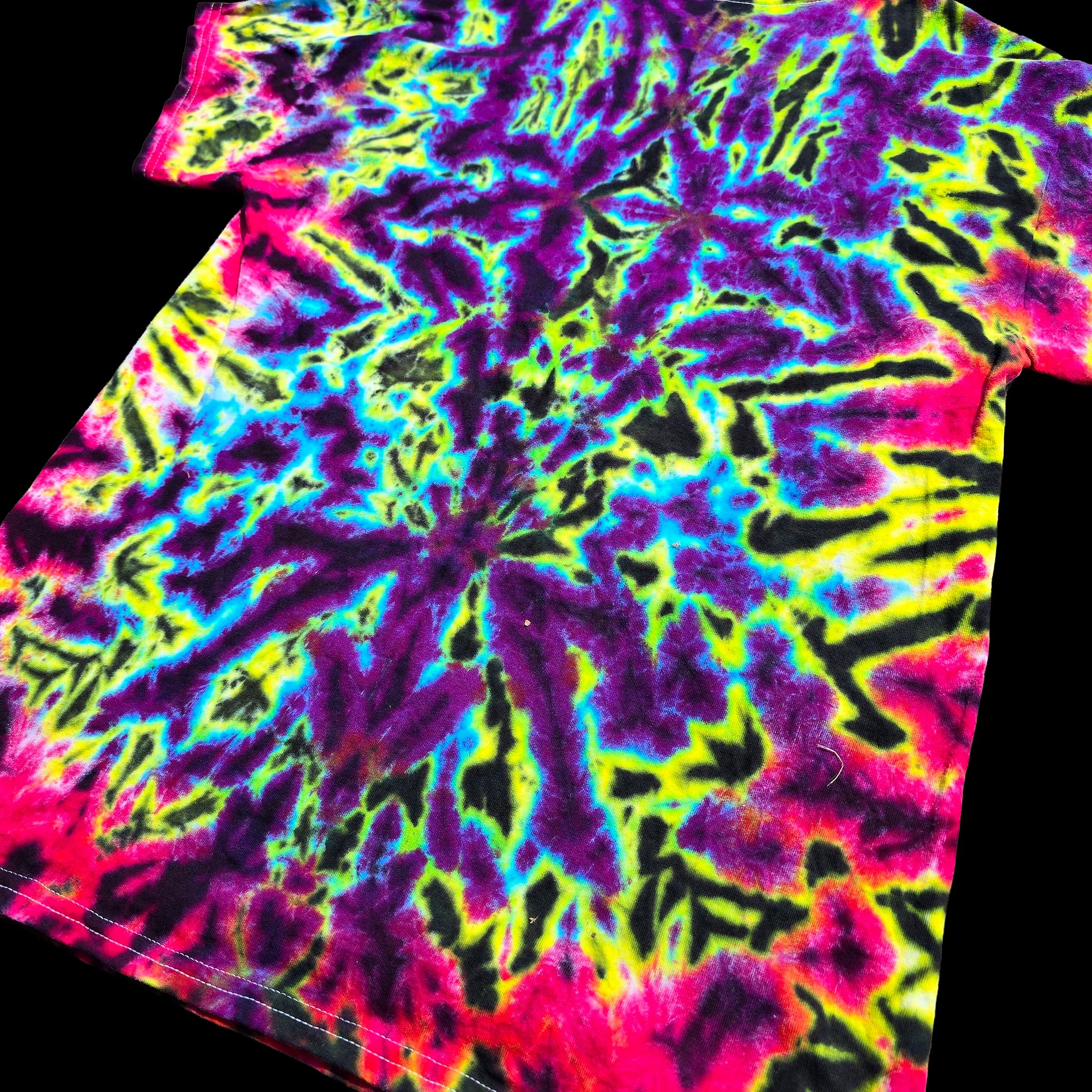 A short-sleeve tie-dye T-shirt laid flat on a black background, featuring a highly saturated neon pattern with purple, pink, yellow, blue, and black in roughly equal prominence.