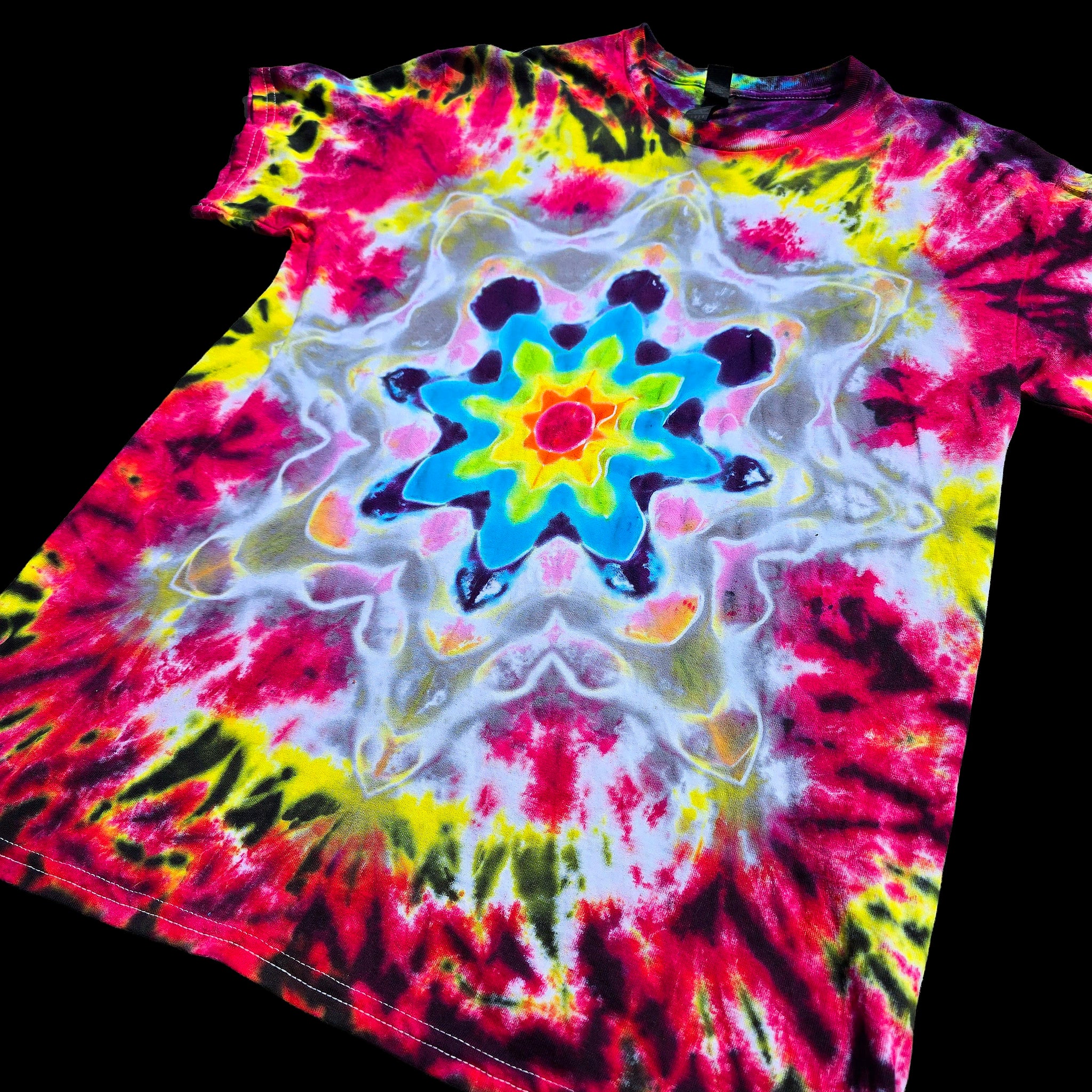 A short-sleeve tie-dye T-shirt laid flat on a black background, featuring a bright central mandala-style floral pattern with red, pink, yellow, blue, white, and black dye blends.