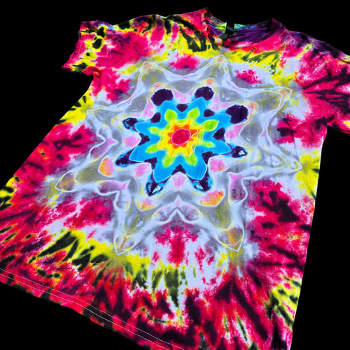 A short-sleeve tie-dye T-shirt laid flat on a black background, featuring a bright central mandala-style floral pattern with red, pink, yellow, blue, white, and black dye blends.