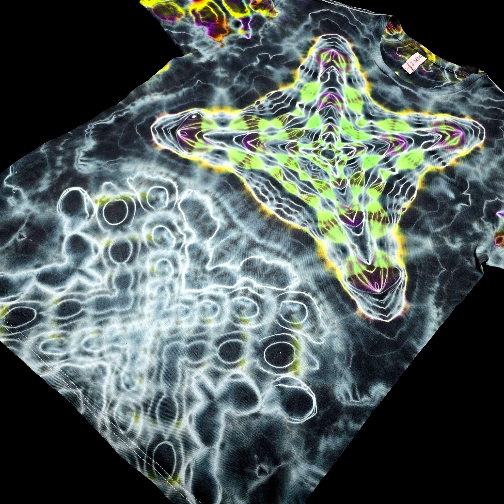 A black T-shirt laid flat on a black background, featuring a large neon green star-like graphic outlined in yellow and purple, with white bubble-like circular patterns across the rest of the shirt.