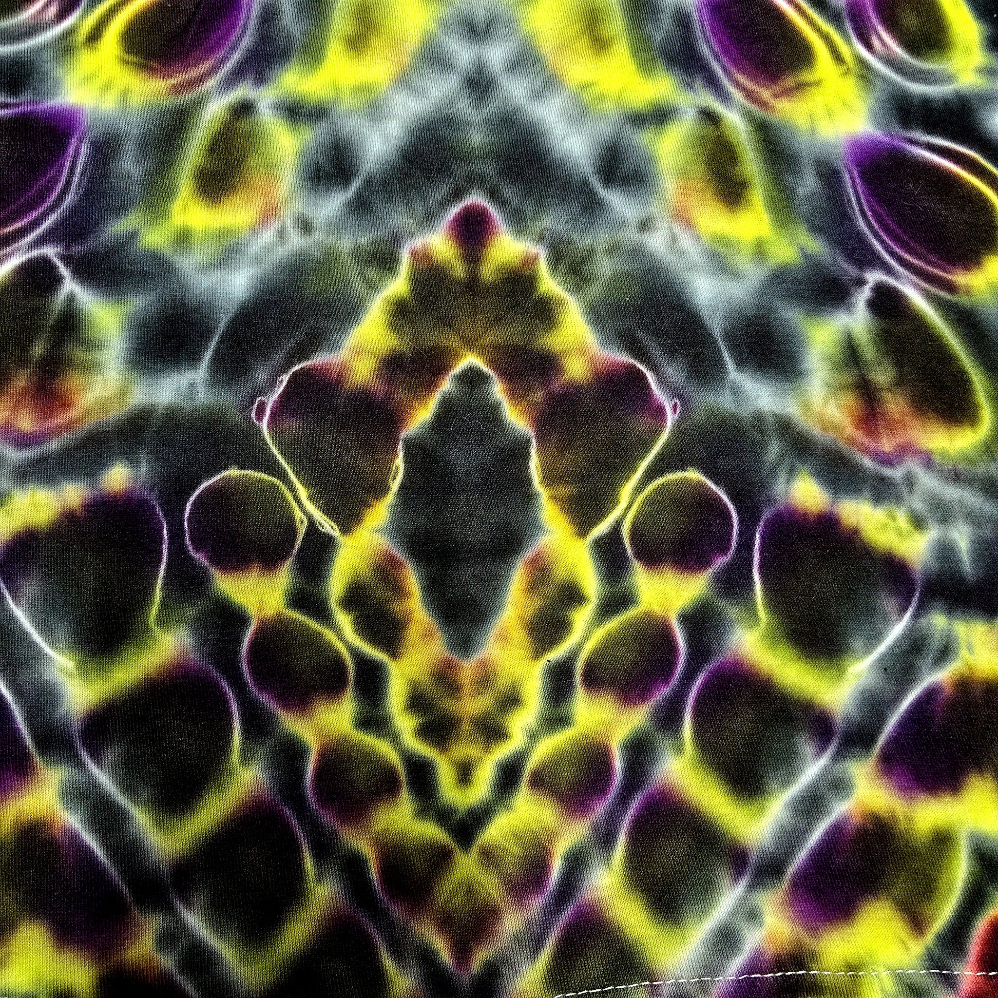A close-up of a tie-dye fabric with a symmetrical, kaleidoscopic pattern in vibrant yellow, purple, black, and gray.