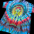 A short-sleeve tie-dye T-shirt laid flat on a black background, featuring a spiral burst pattern with bright blue, red, green, yellow, and black dye blending through the fabric.