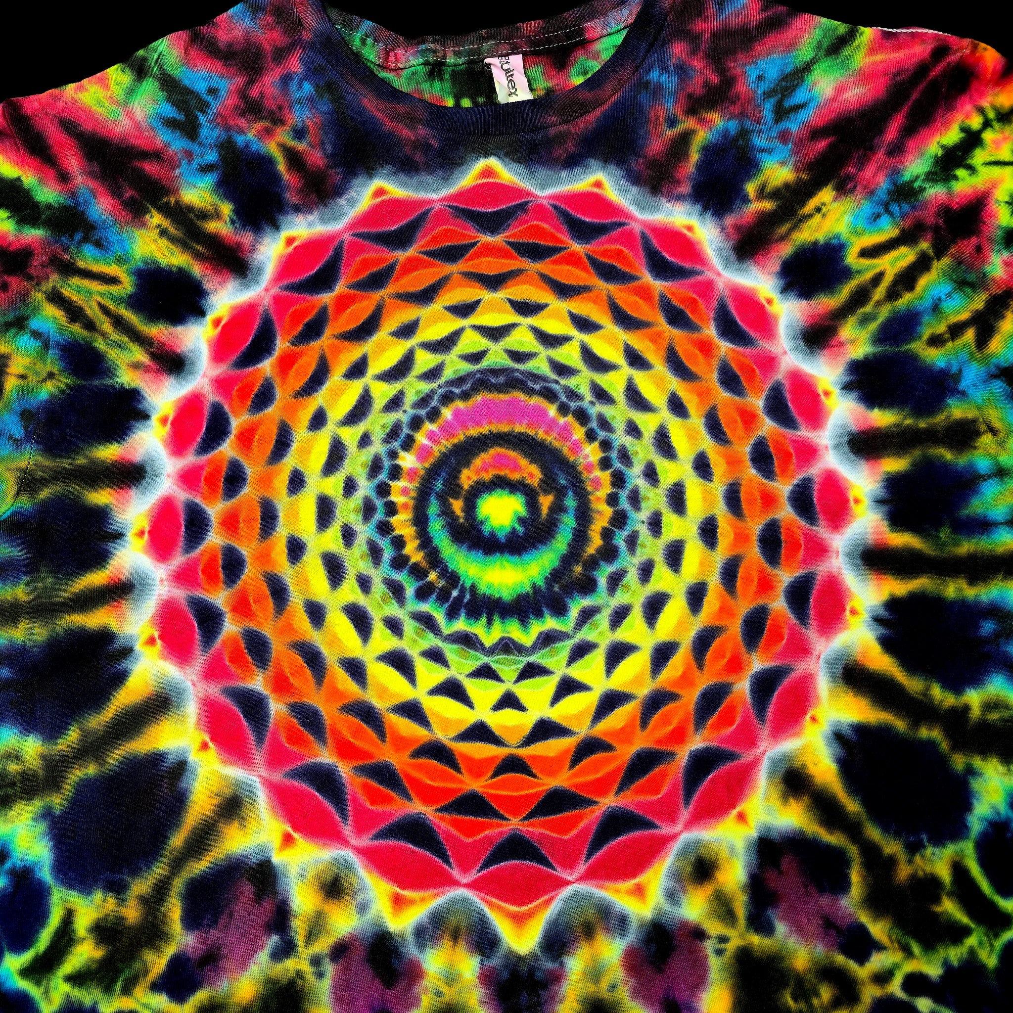 A close-up of a tie-dye t-shirt featuring a vibrant, symmetrical mandala pattern with concentric circles and geometric shapes in neon yellow, orange, red, pink, green, blue, and purple.