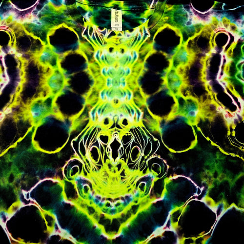 A close-up of a tie-dye t-shirt featuring a highly symmetrical, psychedelic pattern in neon green, lime yellow, black, and purple. A white neck label with the brand name 'DAMAX' is visible at the top center.
