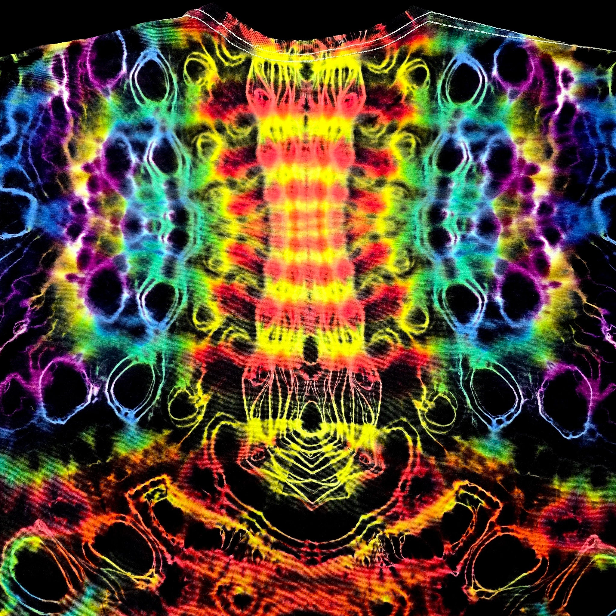 A close-up of a black garment covered in a vivid neon tie-dye pattern with mirrored rainbow stripes and circular cutout shapes in yellow, red, orange, green, blue, and purple.