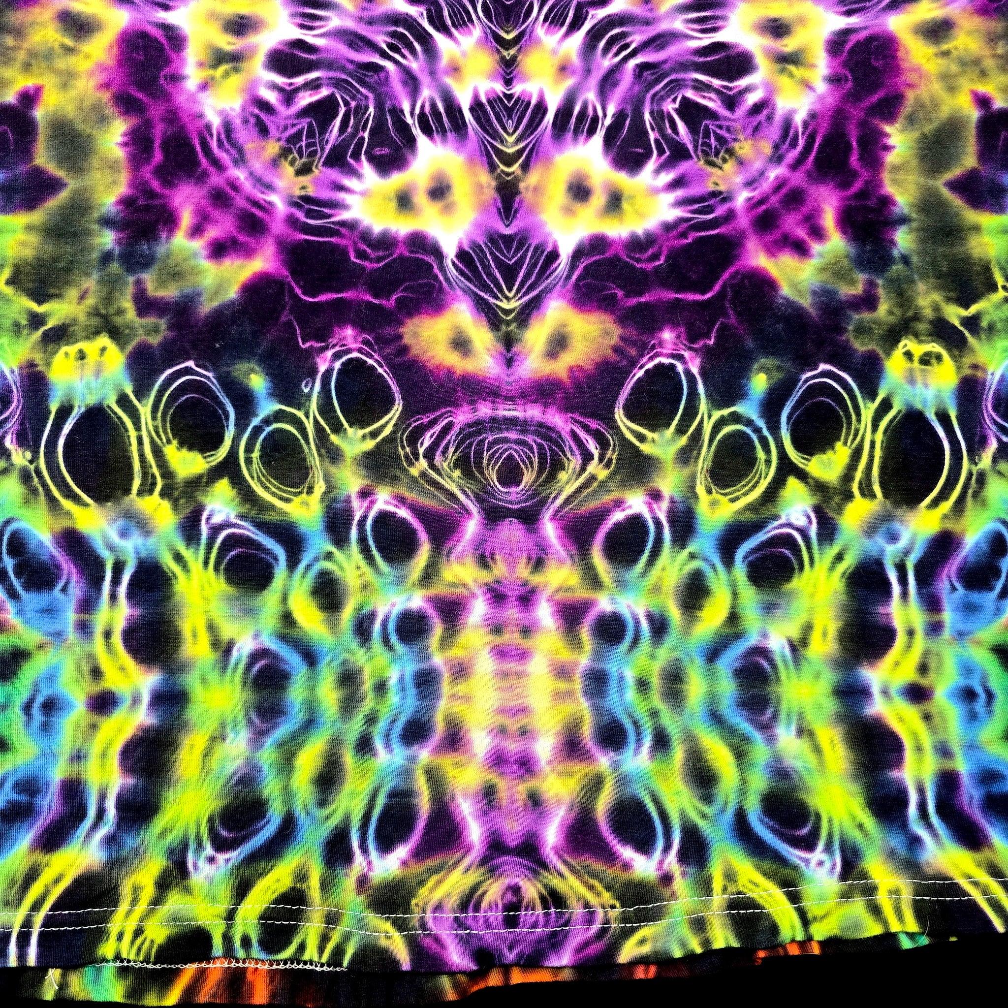 A close-up of a tie-dye fabric with a highly symmetrical, kaleidoscopic pattern in neon purple, yellow, green, blue, and black.