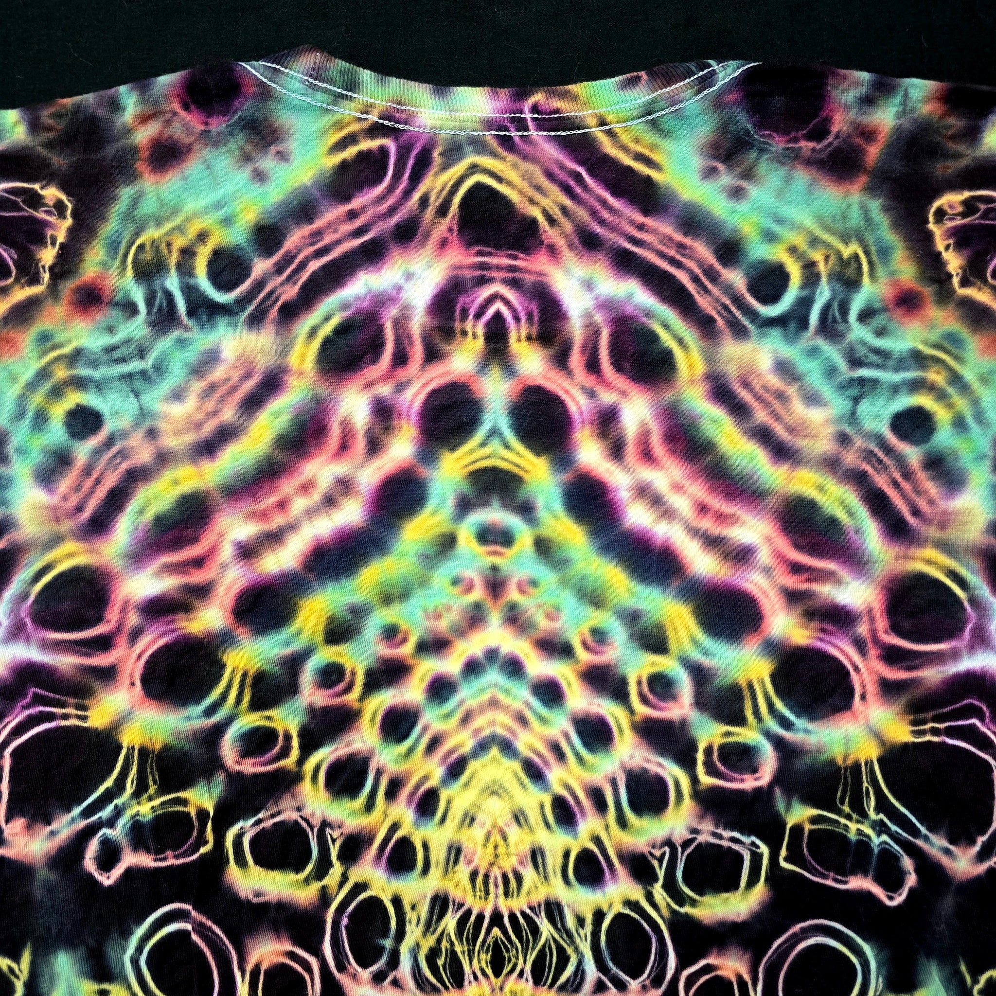 A close-up of a tie-dye t-shirt featuring a highly symmetrical, psychedelic pattern with neon pink, yellow, green, and blue outlines on a black base.